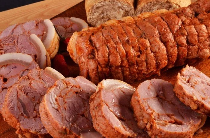 Un arrollado de huaso artesanal, amarrado con cordel y untado en ají rojo, servido en rodajas sobre una tabla de madera.