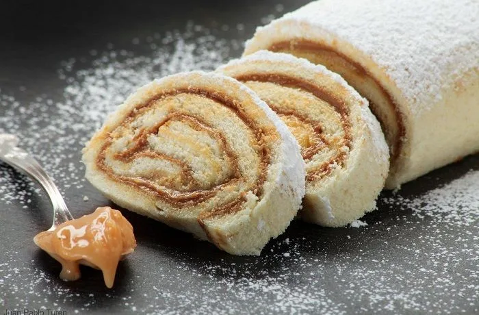 Brazo de reina Brazo de reina chileno espolvoreado con azúcar flor, relleno de abundante manjar sobre un fondo rústico.