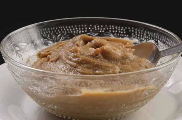 Un pocillo de greda con dulce de harina dorada de color café brillante, de consistencia espesa, servido junto a una cuchara de madera.