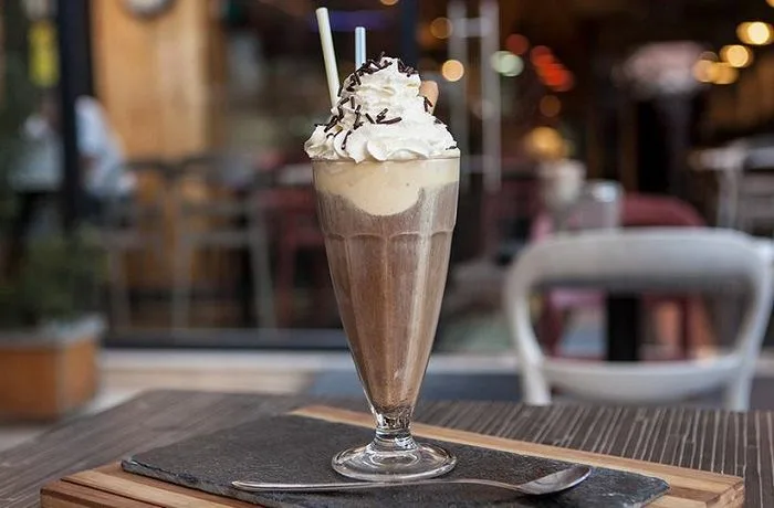 Café helado chileno Un vaso alto de café helado con dos bolas de vainilla, una montaña de crema chantilly, salsa de manjar chorreando y dos galletas wafer decorativas.