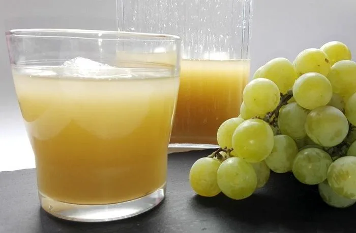 Pronto alivio chileno Una botella de vidrio transparente llena de Pronto Alivio, mostrando su color amarillo pálido, junto a un racimo de uvas blancas frescas.