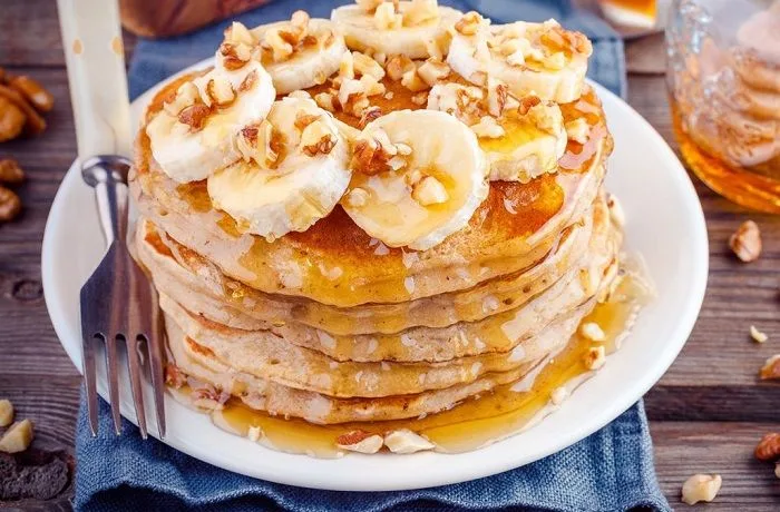 Panqueques de avena y plátano Torre de panqueques de avena y plátano decorados con rodajas de fruta fresca y un chorro de miel líquida sobre un plato artesanal.