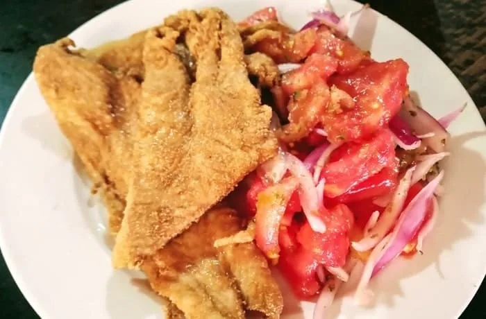 Un plato con filetes de merluza frita de color café dorado, con una textura rugosa gracias a la harina tostada, acompañados de una rodaja de limón.