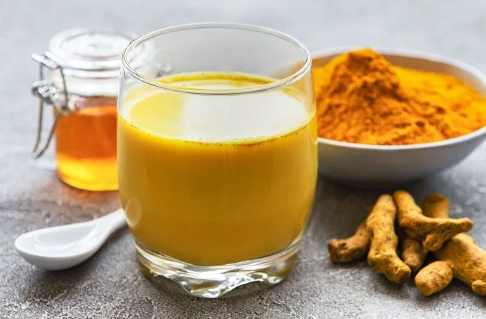 Receta de leche de oro o dorada: La ancestral golden milk hindú 1 Una taza de cristal con leche de oro espumosa, decorada con una rama de canela y espolvoreada con cúrcuma en la superficie.