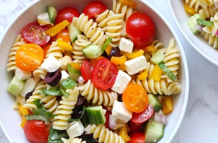 Un bol de ensalada de fideos espirales con hojas verdes, tomates cherry rojos, trozos de mozzarella blanca y pimentón, con un aliño brillante de aceite y hierbas.