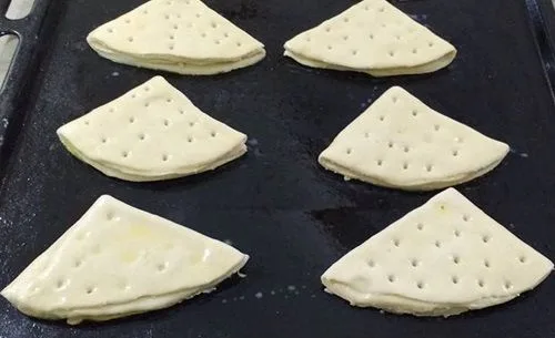 Dobladitas en bandeja de horno Dobladitas dobladas en forma triangular, pinceladas con huevo, sobre una lata de horno.