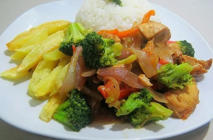 Saltado de brócoli oriental con pollo y vegetales en un plato, acompañado de arroz blanco.