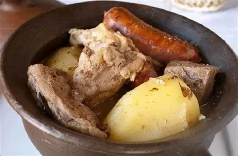 Receta de estofado de San Juan chileno Plato hondo de greda con estofado de San Juan chileno humeante, mostrando presas de pollo dorado, costillar de cerdo ahumado y rodajas de longaniza en caldo de vino.