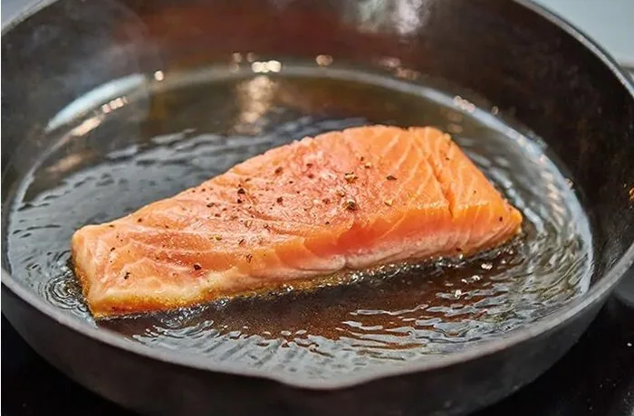 Un filete de salmón perfectamente dorado en una sartén, bañado en mantequilla derretida con hierbas frescas y rodajas de limón al costado.