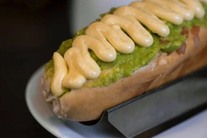 Receta de completos mojados o talquinos Primer plano de dos completos mojados o talquinos, con salchichas, tomate rallado, palta molida y mayonesa casera, en un soporte de hot dog